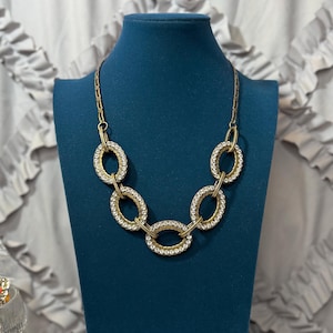 May include: A gold-coloured necklace featuring oval links adorned with small, clear crystals. The necklace is displayed on a dark teal mannequin. The background is a light, ruffled fabric.