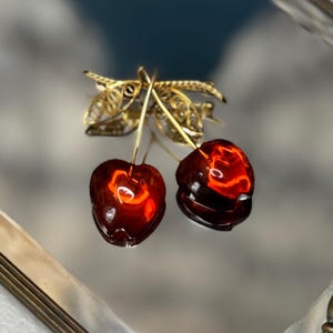 May include: A pair of cherry-shaped earrings with a gold-colored filigree leaf design. The cherries are a deep red with a glowing interior, and the stems are gold. The earrings are displayed on a reflective surface.