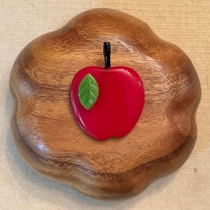 May include: A red apple-shaped brooch with a green leaf and black stem, set on a wooden base. The brooch is a vibrant red color, and the wooden base has a natural wood grain pattern.