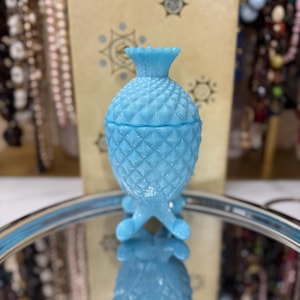 Könnte beinhalten: Eine hellblaue Glasvase in Ananasform mit Deckel und drei Beinen. Die Vase steht auf einem silbernen Tablett mit einer spiegelnden Oberfläche.