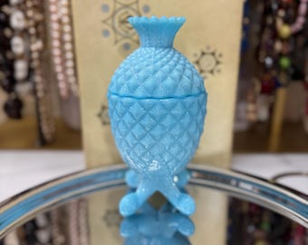 May include: A light blue glass pineapple-shaped jar with a lid and three legs. The jar is sitting on a silver tray with a mirrored surface.