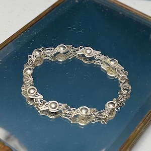 Puede incluir: Un brazalete de filigrana plateada con eslabones ovalados, cada uno con un elemento circular central. El brazalete se muestra sobre una superficie reflectante, destacando su intrincado diseño y delicada artesanía.