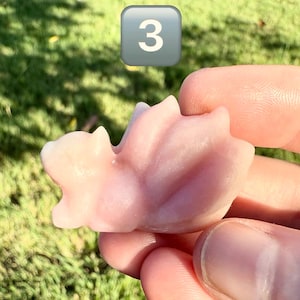 May include: A light pink, carved stone figurine, possibly a small animal or mythical creature, held in a person's hand. The figurine is smooth and has rounded edges. A gray square with the number 3 is in the background.