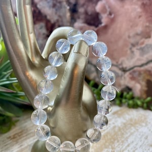 May include: A clear quartz beaded bracelet. The bracelet is composed of round, translucent beads strung together. The beads are displayed on a gold-colored hand-shaped display stand, with a blurred background of pink and green.