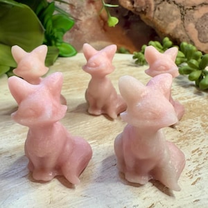 May include: Five pink fox figurines are arranged on a light-colored wooden surface. The foxes are in a sitting position with their ears perked up. The figurines have a smooth, polished appearance and are a soft pink color.