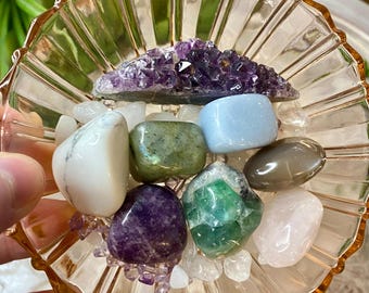 Puede incluir: Una colección de piedras preciosas pulidas en varios colores, incluyendo blanco, verde, azul, morado y rosa, dispuestas en un cuenco de vidrio transparente y acanalado. El cuenco tiene un tono anaranjado rosado y está lleno de pequeños cristales transparentes.