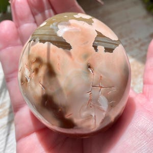 May include: A polished, oval-shaped stone with a blend of colors including peach, brown, and cream. The stone has a smooth, glossy surface with natural patterns and some visible cracks. The stone is held in a hand.