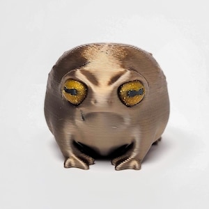 May include: A small, brown, 3D printed frog figurine with yellow eyes. The frog is sitting on a white surface.