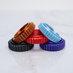 May include: A collection of colorful, textured rings in various shades including brown, red, blue, purple, and black. Each ring features a unique geometric pattern on its outer edge, creating a tactile and visually appealing design. The rings are stacked, showcasing their diverse colors and textures.