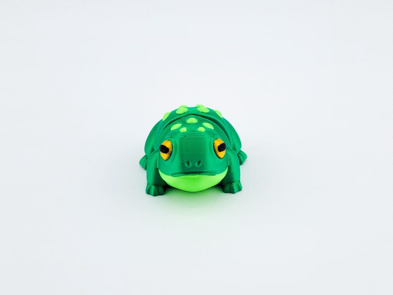 May include: A green 3D printed frog figurine with yellow eyes and a textured surface.