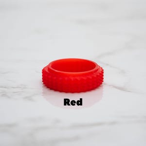 May include: A vibrant red ring-shaped object with a textured, spiky exterior. The word "Red" is printed in black below the object. The ring is set against a white marble-like background.