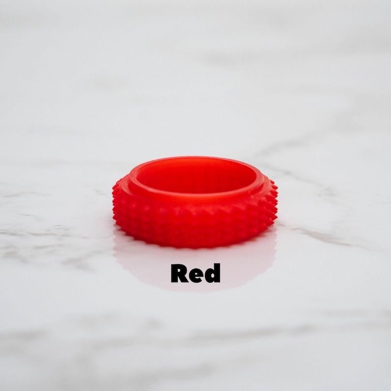 May include: A vibrant red ring-shaped object with a textured, spiky exterior. The word "Red" is printed in black below the object. The ring is set against a white marble-like background.