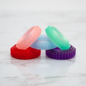 Könnte beinhalten: Eine Ansammlung farbenfroher, strukturierter Ringe in Rot, Rosa, Blau, Grün und Lila. Die Ringe sind auf einer weißen Marmoroberfläche gestapelt und angeordnet.