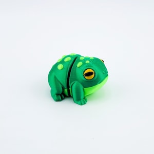 May include: A green 3D printed frog figurine with yellow eyes. The frog is sitting on a white surface.