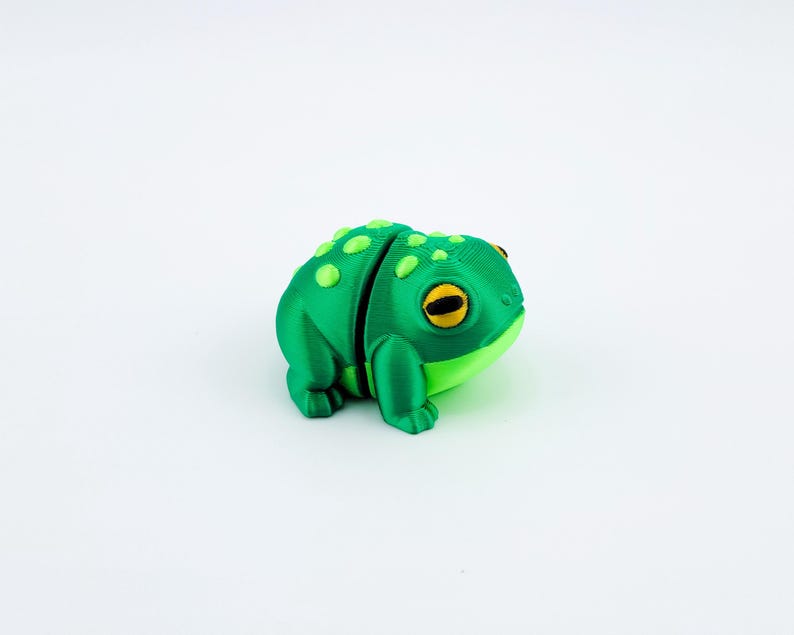 May include: A green 3D printed frog figurine with yellow eyes. The frog is sitting on a white surface.