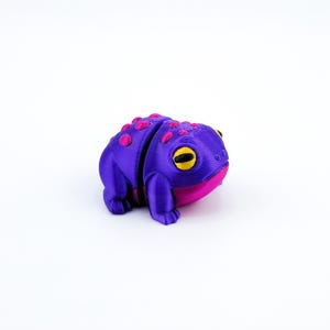 May include: A small, purple and pink 3D printed frog figurine with yellow eyes. The frog is sitting on a white surface.