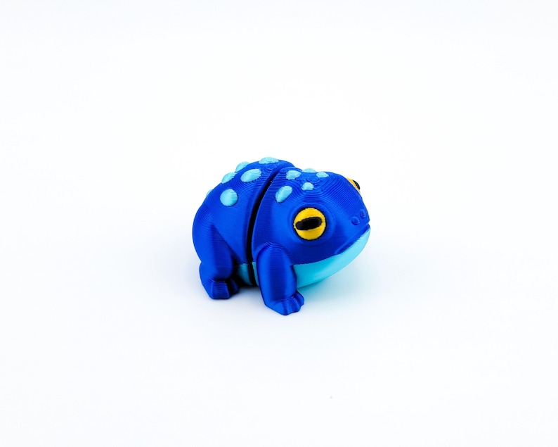 May include: A blue and light blue 3D printed frog figurine with yellow eyes. The frog is sitting on a white surface.