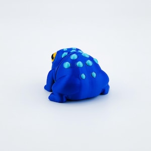 May include: A blue frog figurine with light blue spots. The frog is sitting on a white surface.