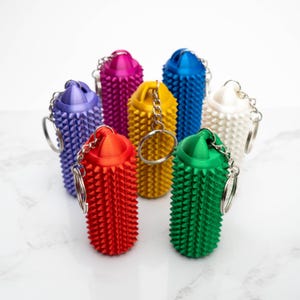 Op de afbeelding: Een verzameling kleurrijke, getextureerde sleutelhangers in verschillende tinten, waaronder rood, geel, groen, blauw, paars, roze en wit. Elke sleutelhanger heeft een cilindrische vorm met een stekelig oppervlak en een zilveren sleutelring.