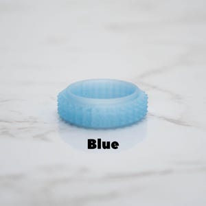 May include: A light blue ring with a textured exterior and the word "Blue" in black text. The ring is circular and appears to be made of a translucent material. The background is a white marble surface.