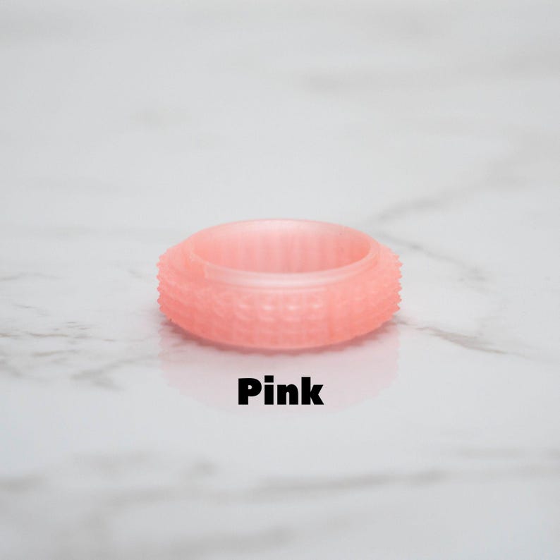 May include: A pink ring with a textured surface and the word "Pink" in black text. The ring has a series of small, raised bumps around its outer edge. The ring is made of a translucent material.