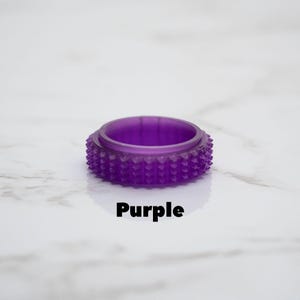 May include: A translucent purple ring with a textured, studded exterior. The word "Purple" is printed in black below the ring. The ring is set against a white marble background.