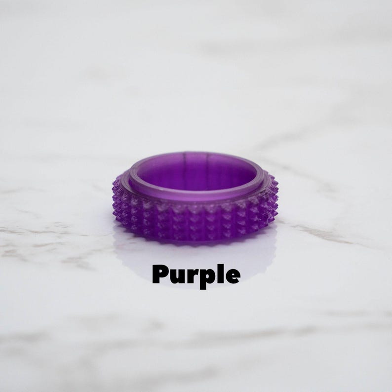 May include: A translucent purple ring with a textured, studded exterior. The word "Purple" is printed in black below the ring. The ring is set against a white marble background.