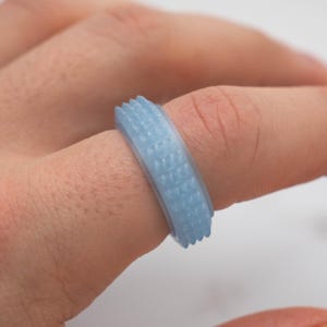 May include: A light blue ring with a textured surface. The ring has a series of small, raised rectangular patterns around the band. The ring is on a finger.