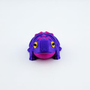 May include: A purple and pink 3D printed frog figurine with yellow eyes. The frog is sitting on a white surface.