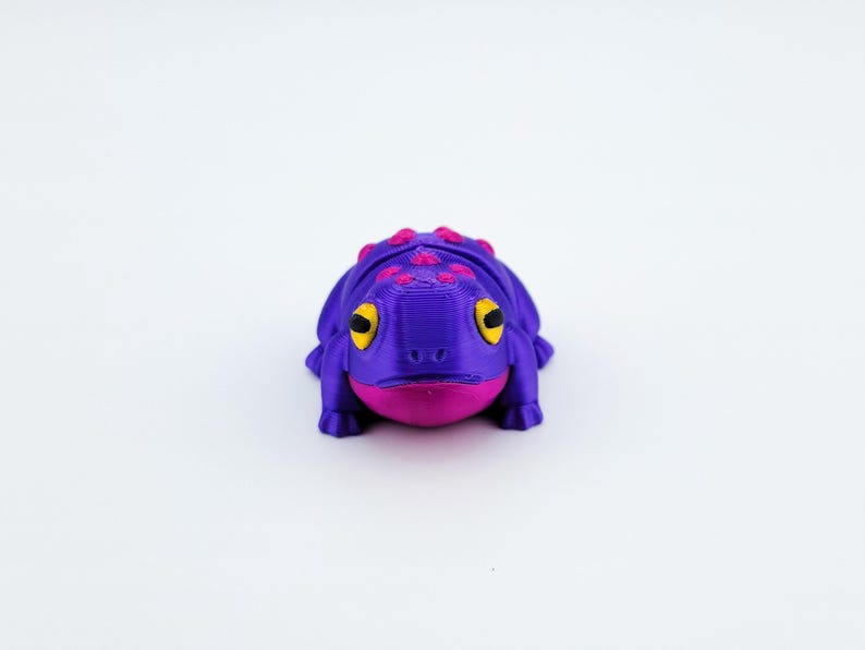 May include: A purple and pink 3D printed frog figurine with yellow eyes. The frog is sitting on a white surface.