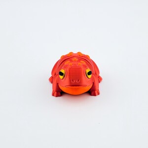May include: A red and orange 3D printed frog figurine with yellow eyes. The frog is sitting on a white surface.