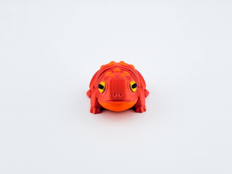 May include: A red and orange 3D printed frog figurine with yellow eyes. The frog is sitting on a white surface.