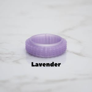 May include: A lavender-colored ring with a textured, studded exterior. The word "Lavender" is printed in black below the ring. The ring is set against a white marble background.