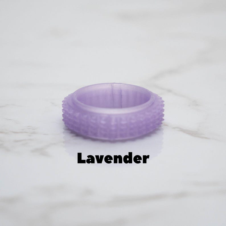May include: A lavender-colored ring with a textured, studded exterior. The word "Lavender" is printed in black below the ring. The ring is set against a white marble background.