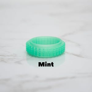 May include: A mint-colored ring with a textured, studded exterior. The ring is a solid, circular band with a slightly raised center. The word "Mint" is printed in black, centered below the ring.