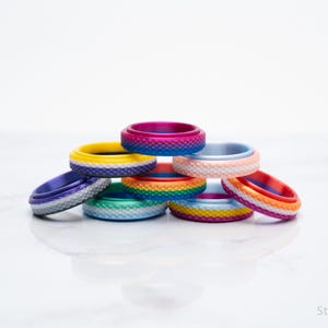 May include: A collection of colorful silicone rings in various shades, including blue, pink, yellow, and orange. The rings are stacked on top of each other, showcasing their textured surfaces and vibrant hues. The rings are a fashion accessory.