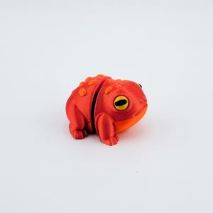 May include: A 3D printed red and orange frog figurine with yellow eyes. The frog is sitting on a white surface.