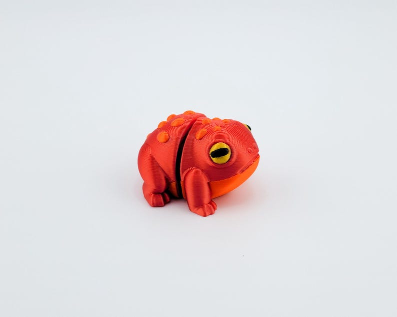 May include: A 3D printed red and orange frog figurine with yellow eyes. The frog is sitting on a white surface.