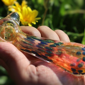 Puede incluir: Una colorida pipa de vidrio con detalles en naranja, azul y verde. La pipa se sostiene en una mano, con un fondo borroso de follaje verde y flores amarillas. El vidrio es transparente.