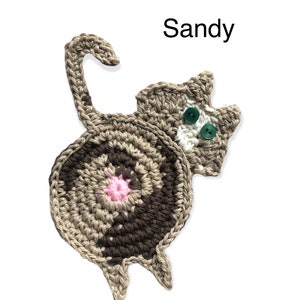 May include: Crocheted cat-shaped coaster in shades of brown, tan, and white, with a pink center and green button eyes. The word "Sandy" is written above the cat. This handmade item is perfect for cat lovers.