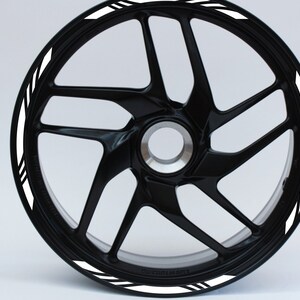 Op de afbeelding: Zwart motorwiel met een multi-spaaks ontwerp. Het wiel heeft een witte streep met zwarte accentlijnen rond de rand. Het wiel staat tegen een effen witte achtergrond.