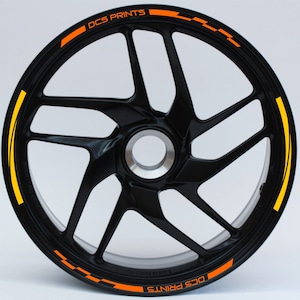 Peut inclure: Roue de moto noire avec des détails orange et jaune. La roue présente un design à plusieurs rayons et le texte "DCS PRINTS" est visible en orange. La jante de la roue présente des rayures orange et jaune.