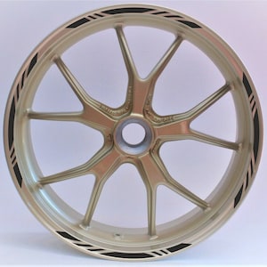 Peut inclure: Une jante de roue de moto dorée avec un design de bande de course noire. La bande est composée de plusieurs fines lignes noires.