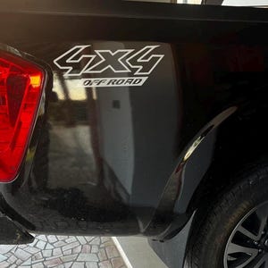 Può includere: Pick-up nero con fanale posteriore rosso e adesivo 4x4 Off Road. Il camion ha una ruota nera con un design a raggi grigio scuro. Il veicolo è parcheggiato su una superficie in pietra.