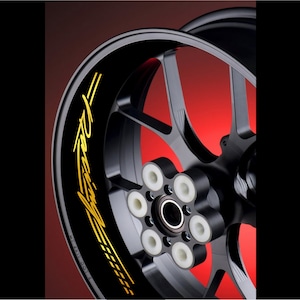Könnte beinhalten: Eine schwarze Motorrad-Felge mit einem goldenen "Racing"-Aufkleber. Der Aufkleber ist um die Felge gebogen und hat ein stilisiertes Rennstreifendesign.