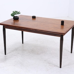 Pode incluir: Mesa de jantar retangular de madeira com pernas castanhas escuras e tampo a condizer. A mesa tem um acabamento natural de grão de madeira. Dois objetos cilíndricos pretos estão sobre a mesa. Um pequeno bonsai e um suporte decorativo também são visíveis.