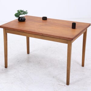 Puede incluir: Una mesa rectangular de madera con cuatro patas, con una tapa marrón claro y un borde marrón más oscuro. Dos pequeños objetos cilíndricos negros están colocados sobre la mesa. También se ven un pequeño bonsái en una maceta negra y una planta decorativa.
