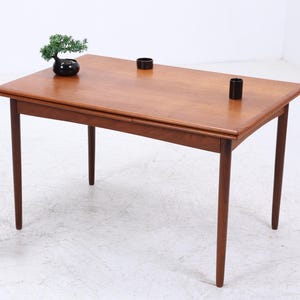 Op de afbeelding: Een rechthoekige houten eettafel met vier poten. De tafel heeft een warme bruine kleur en een glad oppervlak. Twee kleine zwarte cilindrische objecten staan op de tafel. Een kleine bonsai in een zwarte pot staat op de tafel. Een plant en een vaas staan op een houten standaard.