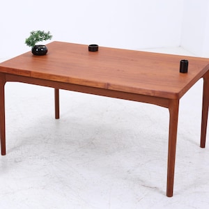 Könnte beinhalten: Ein rechteckiger Holztisch mit warmbraunem Finish. Der Tisch hat vier schlanke Beine und eine glatte Oberfläche. Zwei kleine schwarze zylindrische Objekte sind auf dem Tisch platziert. Eine kleine Pflanze und eine dekorative Vase stehen auf einem Holzständer.