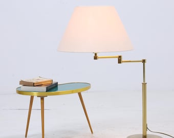 Könnte beinhalten: Messing-Stehleuchte im Mid-Century-Stil mit verstellbarem Arm und cremefarbenem Lampenschirm. Ein kleiner, ovaler Beistelltisch mit türkisfarbener Platte und Holzbeinen steht neben der Lampe. Bücher sind auf dem Tisch gestapelt.